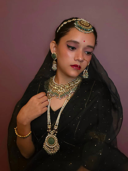 A woman wearing traditional Indian jewelry, including a gold-toned maang tikka, earrings, and a long necklace with green stones, styled with a black embroidered saree against a solid mauve background.
